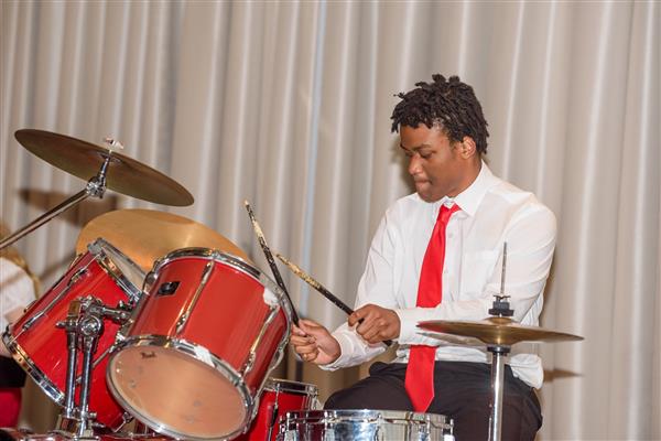 A student in a white button up and a red tie plays the drums while on stage.